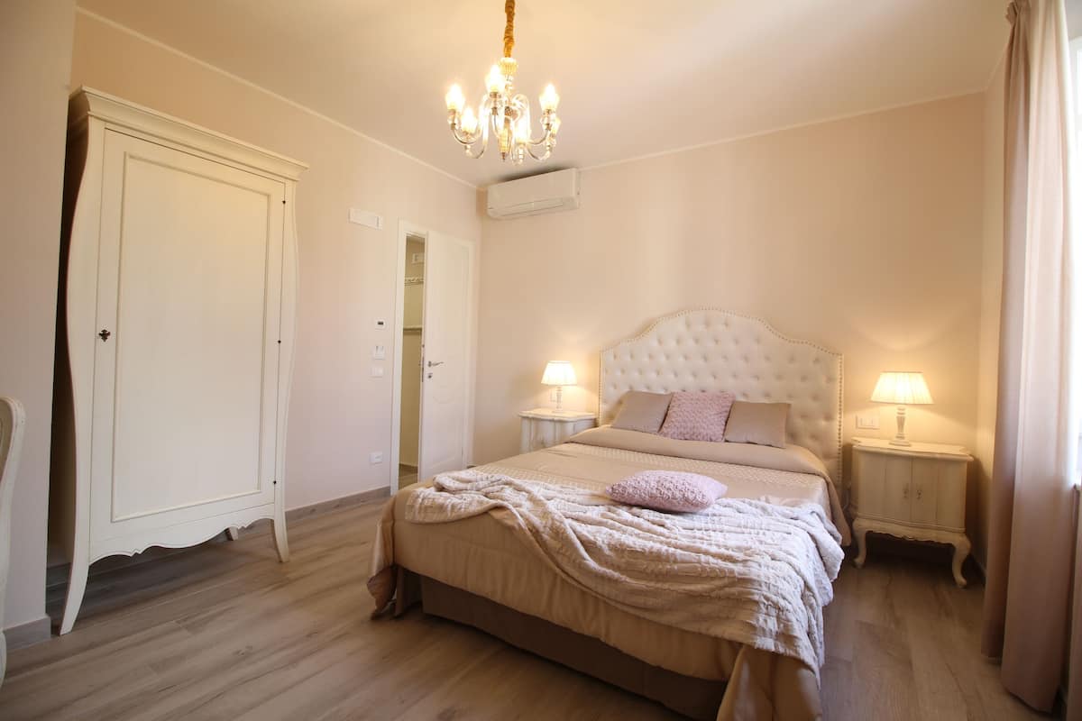 A bedroom with a large bed, two bedside tables with lamps, a chandelier, a wardrobe, and an air conditioning unit.