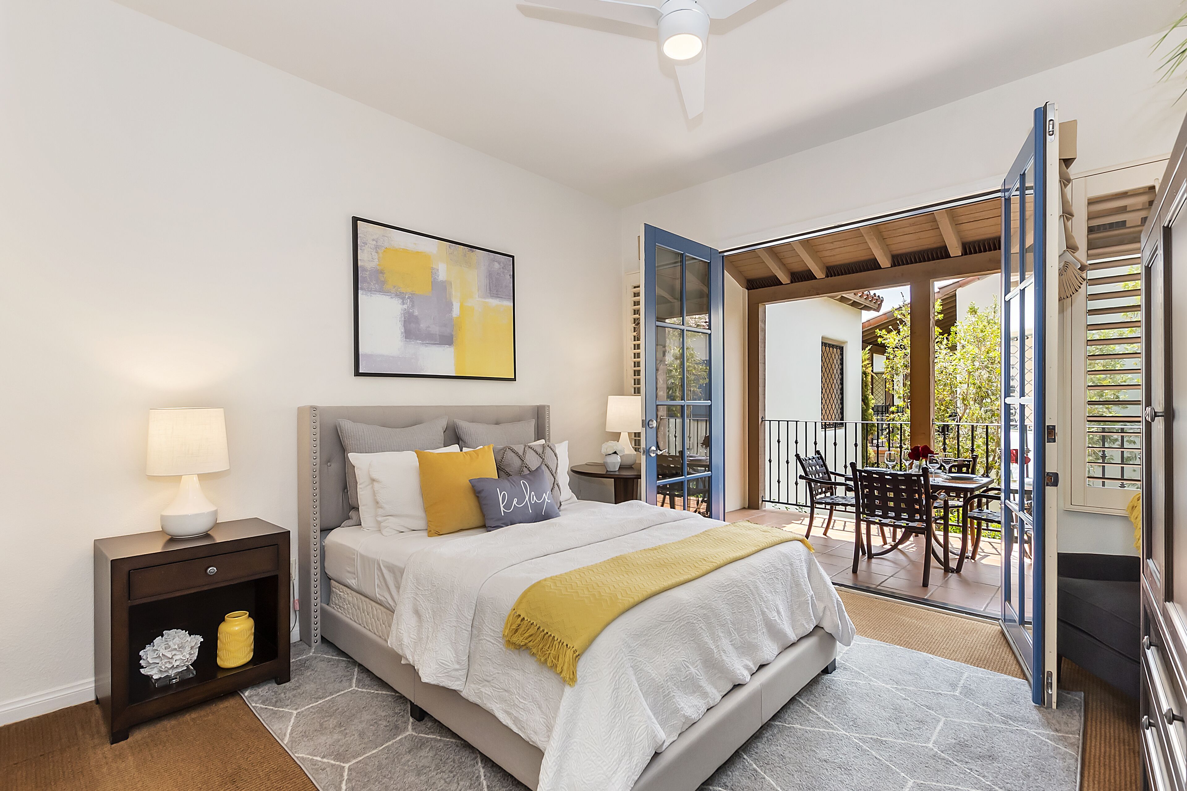A bedroom with a bed, nightstands, a painting, and an open door leading to a balcony with a table and chairs.