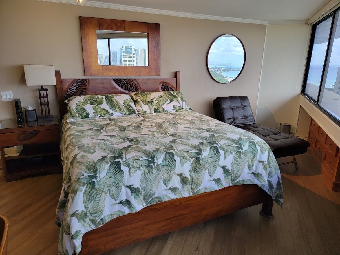 A hotel room with a large bed, a round mirror, a view of the city, and a wooden nightstand with a lamp.