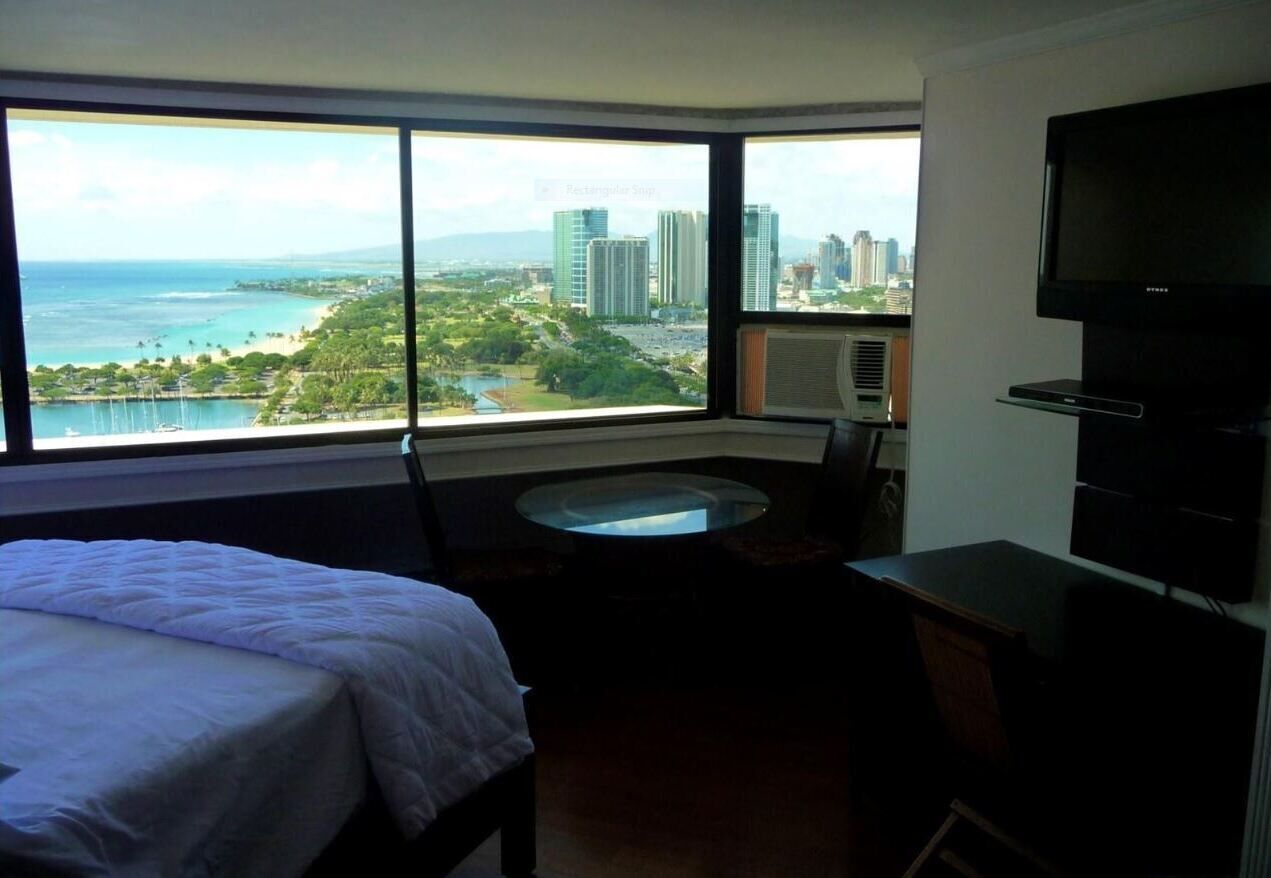 Quarto de hotel com cama, escrivaninha, cadeira, TV e uma grande janela com vista para a paisagem urbana e o oceano.
