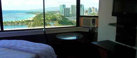 Quarto de hotel com cama, escrivaninha, cadeira, TV e uma grande janela com vista para a paisagem urbana e o oceano.