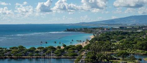 Una marina con numerosas embarcaciones amarradas, una playa con palmeras y un paisaje urbano al fondo.