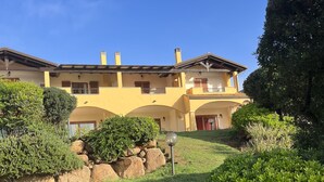 Ein zweistöckiges Gebäude mit gelber Fassade, mehreren Balkonen und einem gepflegten Garten mit Sträuchern und Rasen.