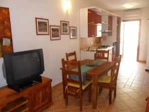 A dining area with a wooden table and chairs, a television on a wooden stand, and a kitchen in the background.
