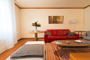 Una sala de estar con un sofá rojo, una mesa redonda de centro, una mesita con un jarrón de flores y un cuadro en la pared.