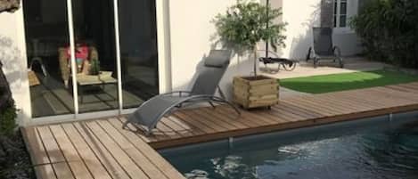 A modern house with a swimming pool, wooden deck, and a sliding glass door leading to a patio area.