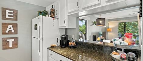 A kitchen with white cabinets, a granite countertop, a coffee maker, and a coffee station.