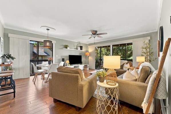 A spacious living room with a sofa, a coffee table, and a TV. There is a dining area with a table and chairs. The room has large windows and a ceiling fan.