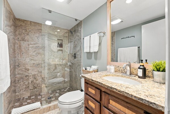 A modern bathroom with a glass-enclosed shower, a sink with a granite countertop, and wooden cabinets. - Beautiful Renovated Pinehurst Condo I 2 Bedroom/ 2 Bath I POOL I sleeps 4 (Pinehurst)