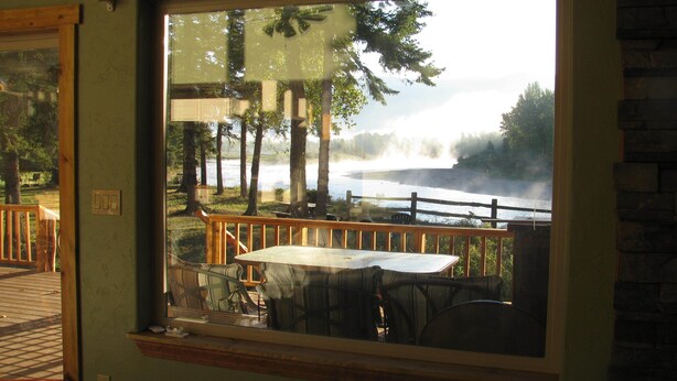 Une vue depuis une fenêtre donnant sur une terrasse avec une table et des chaises, une balustrade en bois et un plan d’eau bordé d’arbres, le tout baigné d’une atmosphère brumeuse.