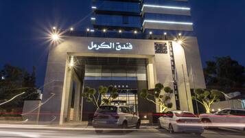 A hotel entrance with a modern facade, illuminated signage, and parked cars in front.