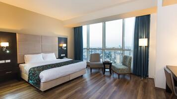 A hotel room with a large bed, a chair, a desk, and a view of the city through large windows.