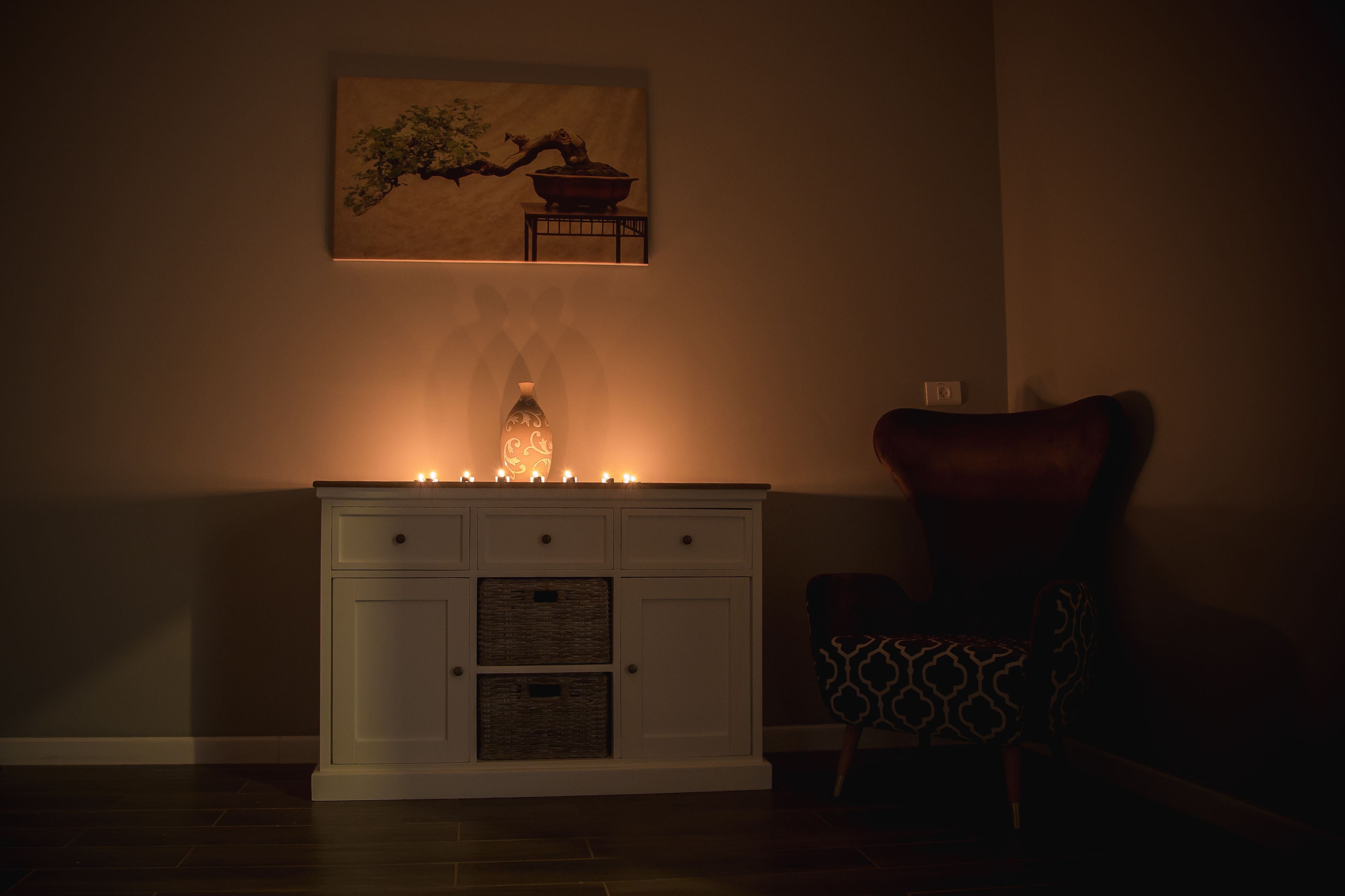 Una stanza scarsamente illuminata con un armadio bianco, una candela accesa, una sedia decorata e un quadro appeso al muro.