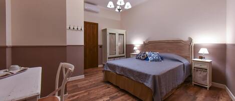 Une chambre avec un plancher en bois, un lit recouvert d’une courtepointe bleue, une table de chevet avec une lampe, une armoire et un bureau avec une chaise.