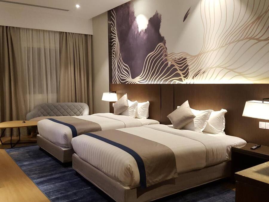 A hotel room with two beds, a desk, a chair, and a large abstract wall art piece.