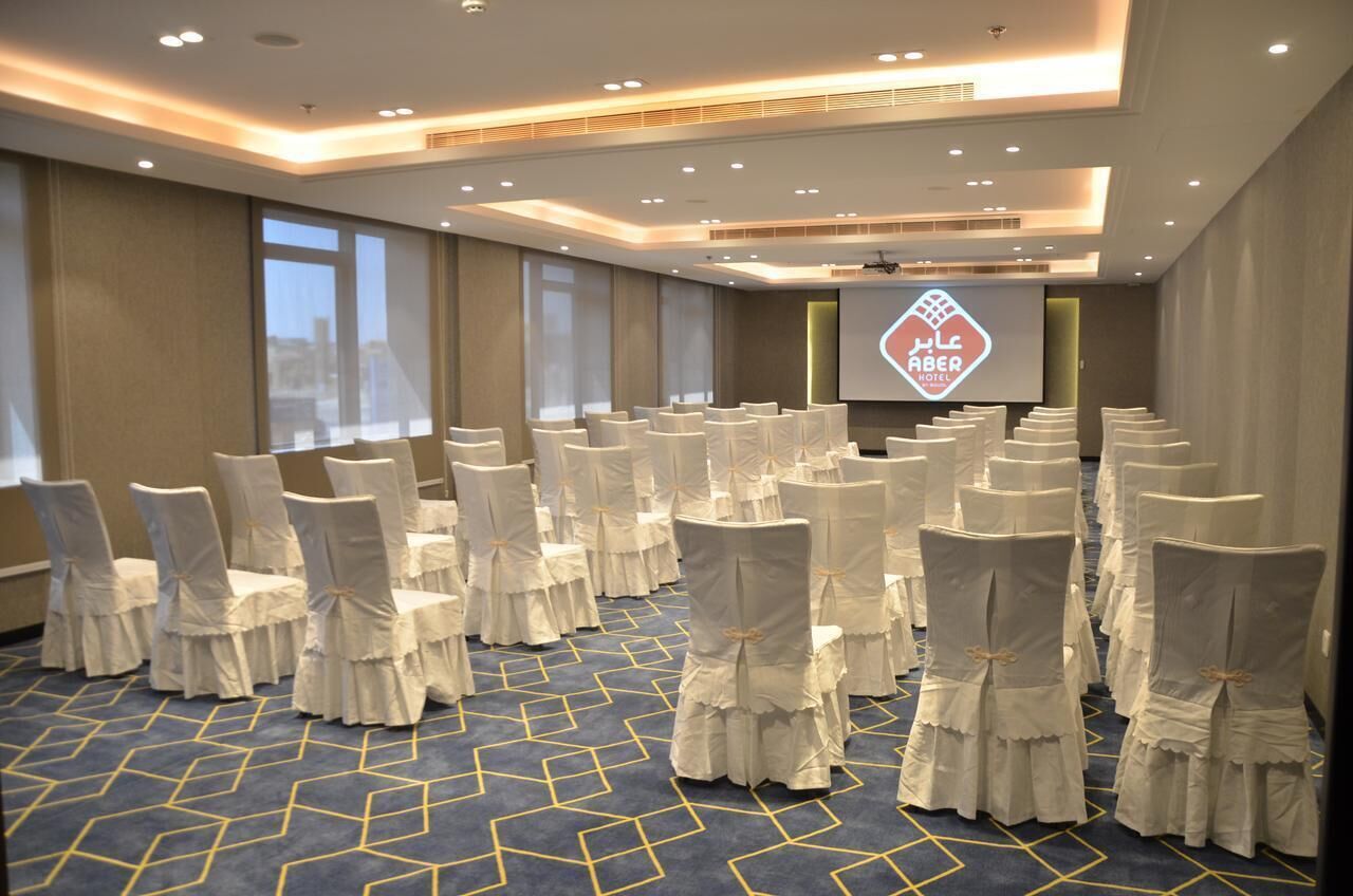 A conference room with rows of chairs covered in white fabric, a large screen displaying a logo, and a patterned carpet.