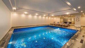Piscina interna moderna com piso de mosaico azul, cercada por cadeiras de lounge e iluminação embutida.