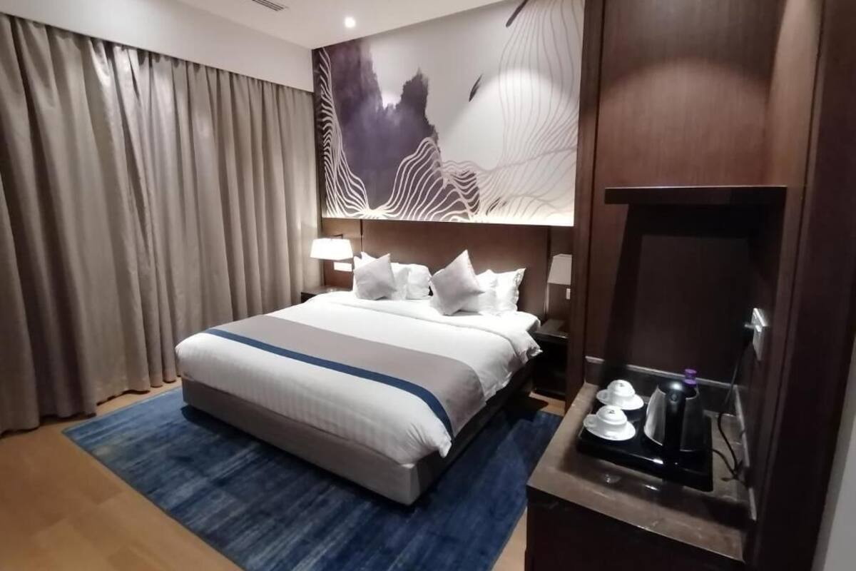 Habitación de hotel con una cama grande, una alfombra azul, una obra de arte en la pared y una mesita con una tetera y tazas.