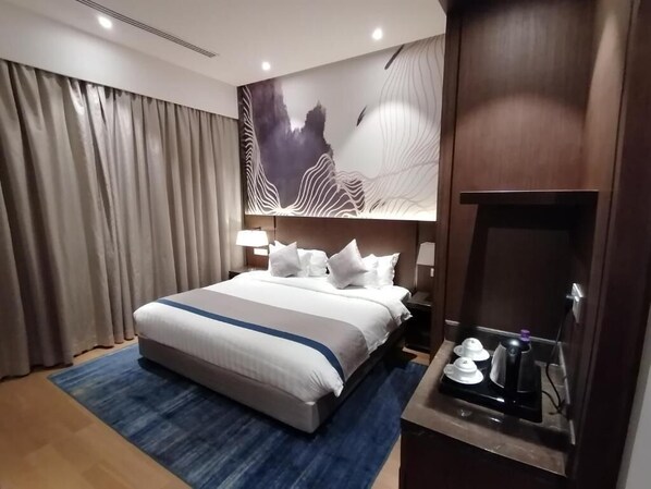 A hotel room with a large bed, a blue rug, a wall art piece, and a small table with a kettle and cups. - Aber Al Yasmin (Riyadh)