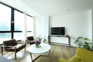 Een moderne woonkamer met een grote televisie, twee leren fauteuils, een ronde salontafel met bloemen en een gele fauteuil.