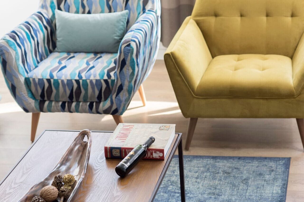 Une pièce avec un fauteuil à rayures, un fauteuil jaune, une table basse en bois sur laquelle se trouvent un livre et une télécommande, et un tapis bleu.