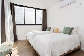 Quarto de hotel com duas camas, janela com cortinas, uma pequena mesa e um aparelho de ar condicionado.