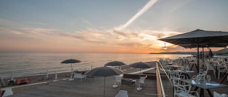 A wooden deck with outdoor seating and umbrellas overlooking the sea at sunset.