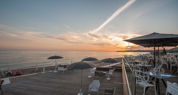 Una terraza de madera con mobiliario exterior y sombrillas, con vistas al mar al atardecer.