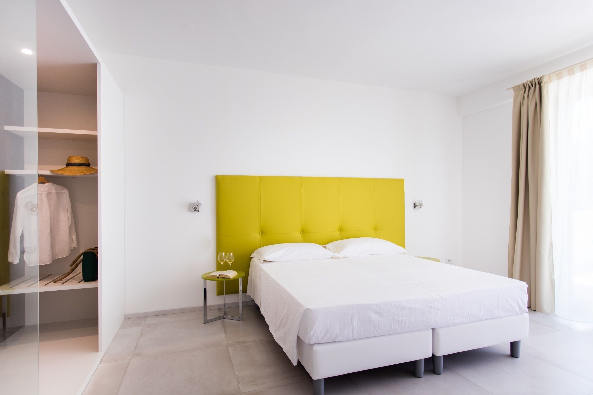 Una habitación de hotel moderna con un cabecero amarillo brillante, una cama bien hecha, una mesita de noche con una copa de vino y un libro, y un armario con ropa colgada.