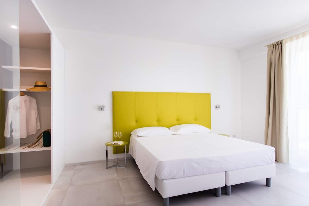 Une chambre d’hôtel moderne avec une tête de lit jaune vif, un lit parfaitement fait, une petite table de chevet avec un verre de vin et un livre, et une armoire contenant des vêtements suspendus.