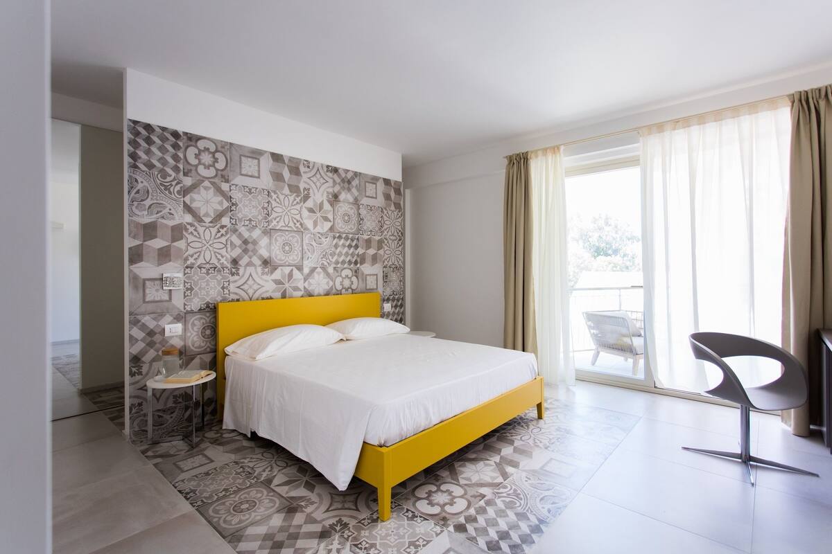 Una habitación de hotel moderna con una cama amarilla, azulejos con diseño en la pared, una mesita auxiliar y una silla junto a la ventana.