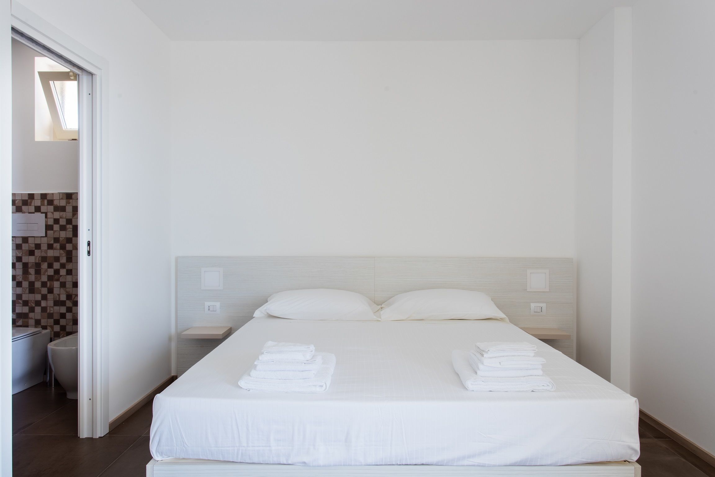 Habitación de hotel limpia y moderna con una cama bien hecha, una mesita de noche pequeña y vistas al baño por una puerta abierta.