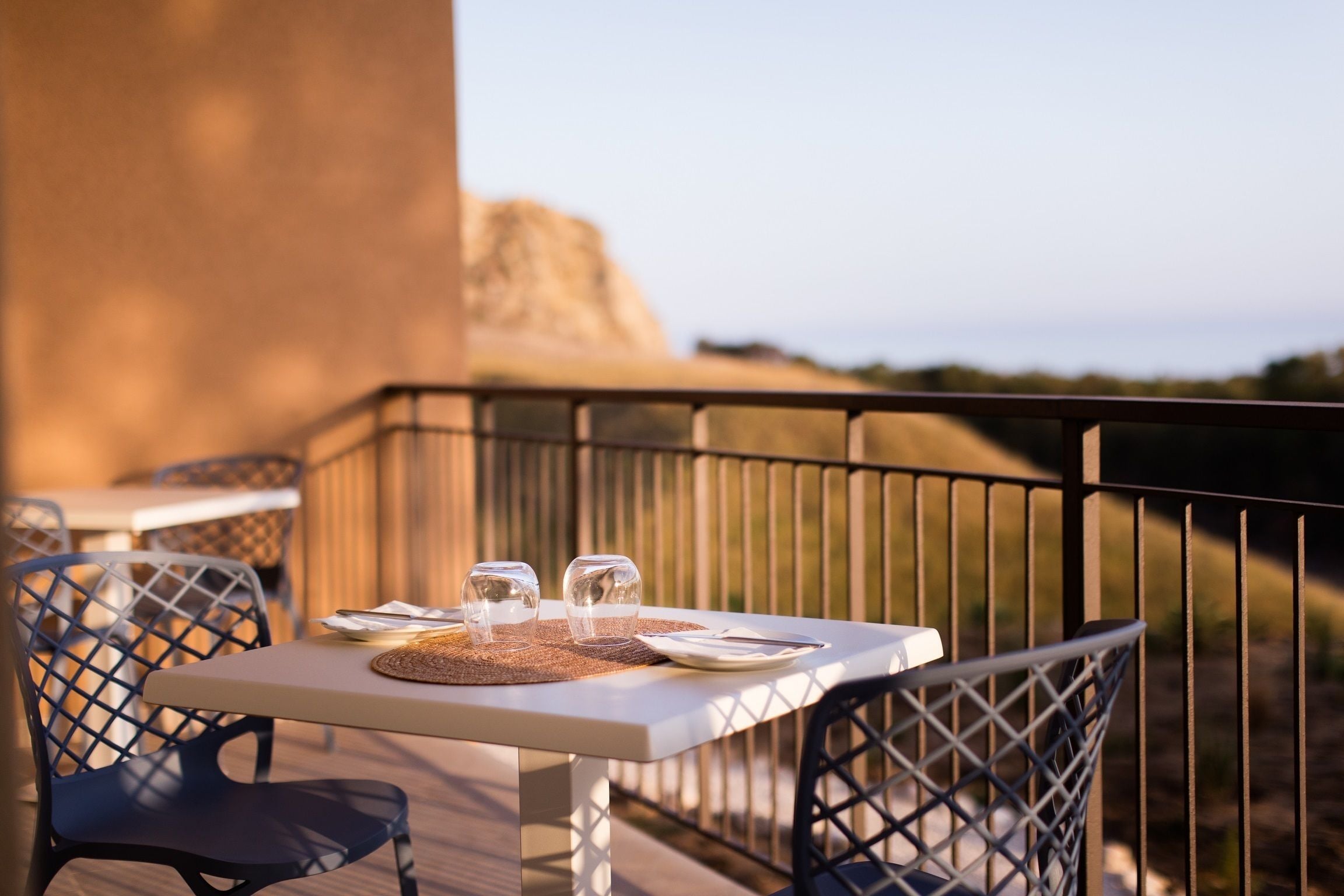 Una mesa dispuesta con vasos y cubiertos en un balcón con sillas de metal, con vistas a un paisaje que incluye una gran formación rocosa.