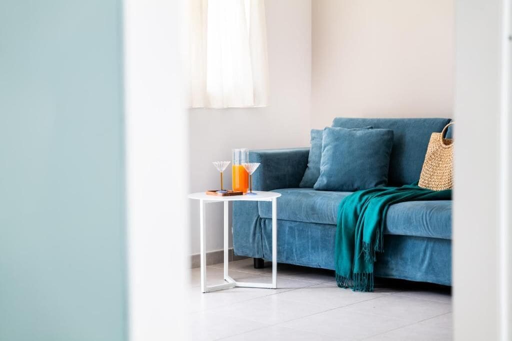 Una habitación con un sofá azul, una mesita blanca con un vaso de zumo de naranja y un bolso tejido.
