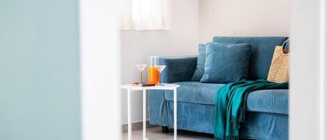 Una habitación con un sofá azul, una mesita blanca con un vaso de zumo de naranja y un bolso tejido.