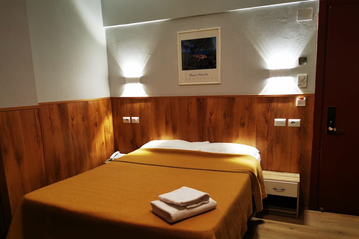 A hotel room with a wooden headboard, a bed with a white comforter, a folded towel on the bed, and a nightstand with a phone.
