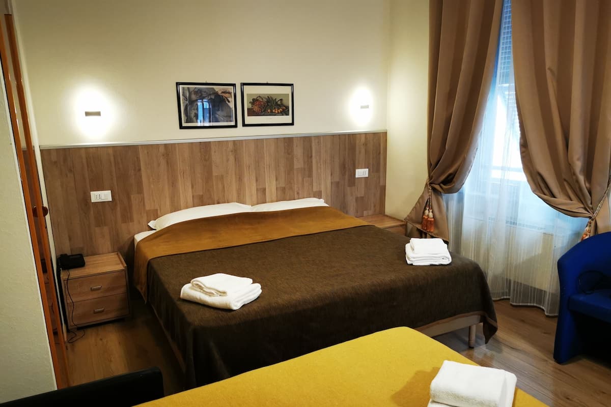 A hotel room with a wooden headboard, two framed pictures on the wall, a bed with a brown bedspread, a yellow table, and a blue chair.
