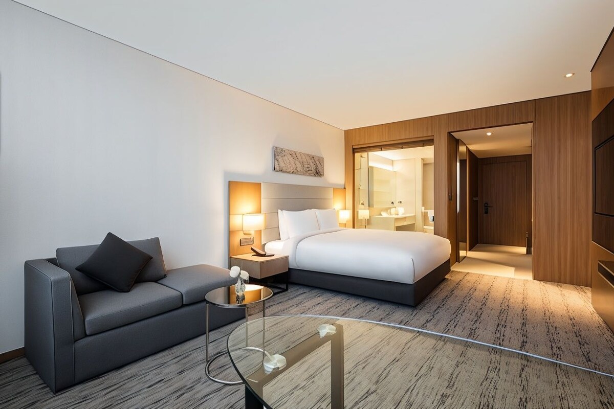 Una habitación de hotel moderna con cama, sofá, mesa de centro de vidrio y mesita de noche con lámparas.