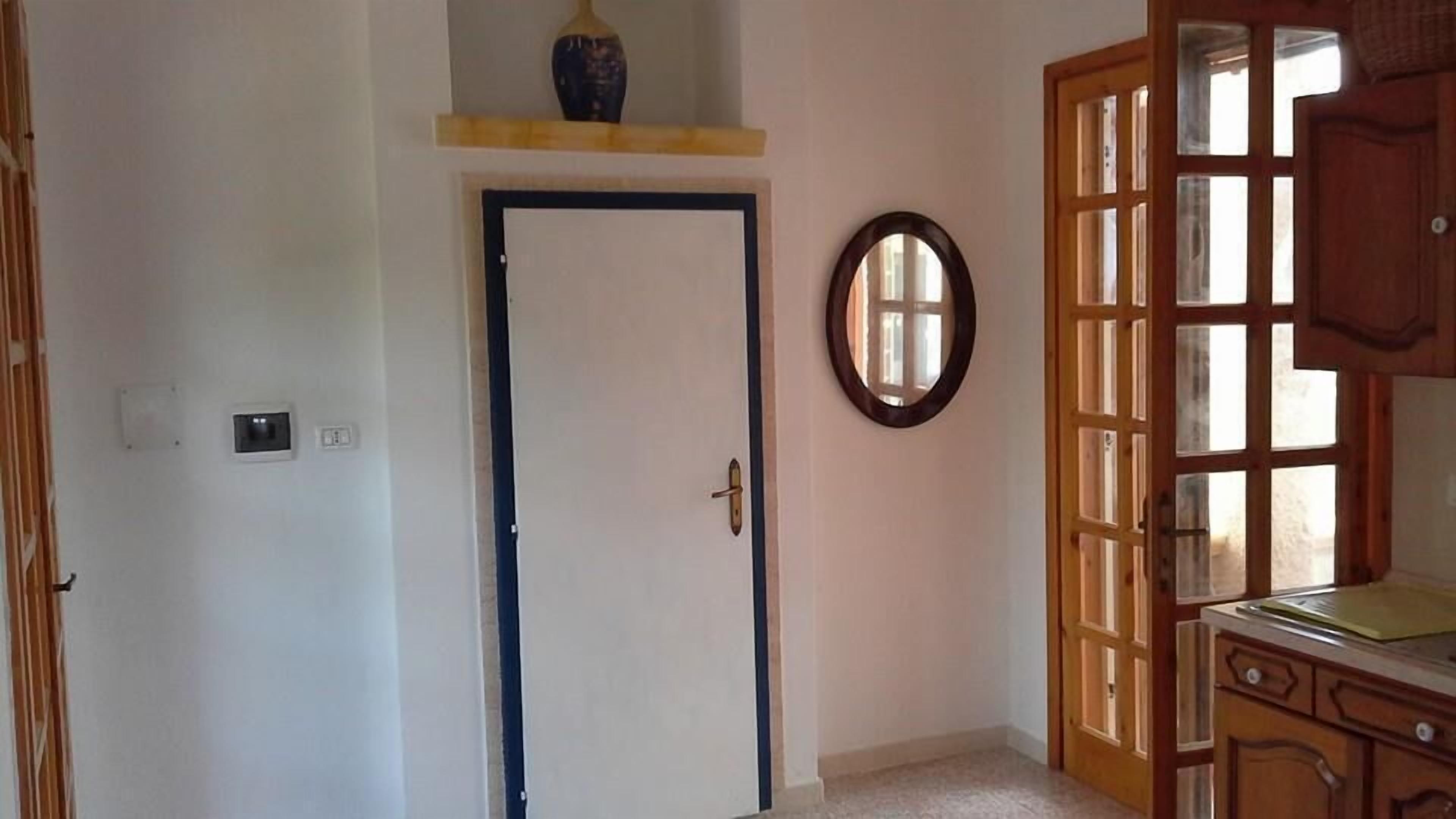 Une pièce avec une porte fermée, une petite étagère sur laquelle se trouve un vase, un miroir au mur et une commode en bois avec des tiroirs.