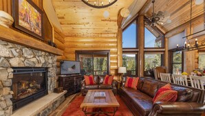A cozy log cabin interior with a stone fireplace, leather sofa, and large windows offering a view of the forest.