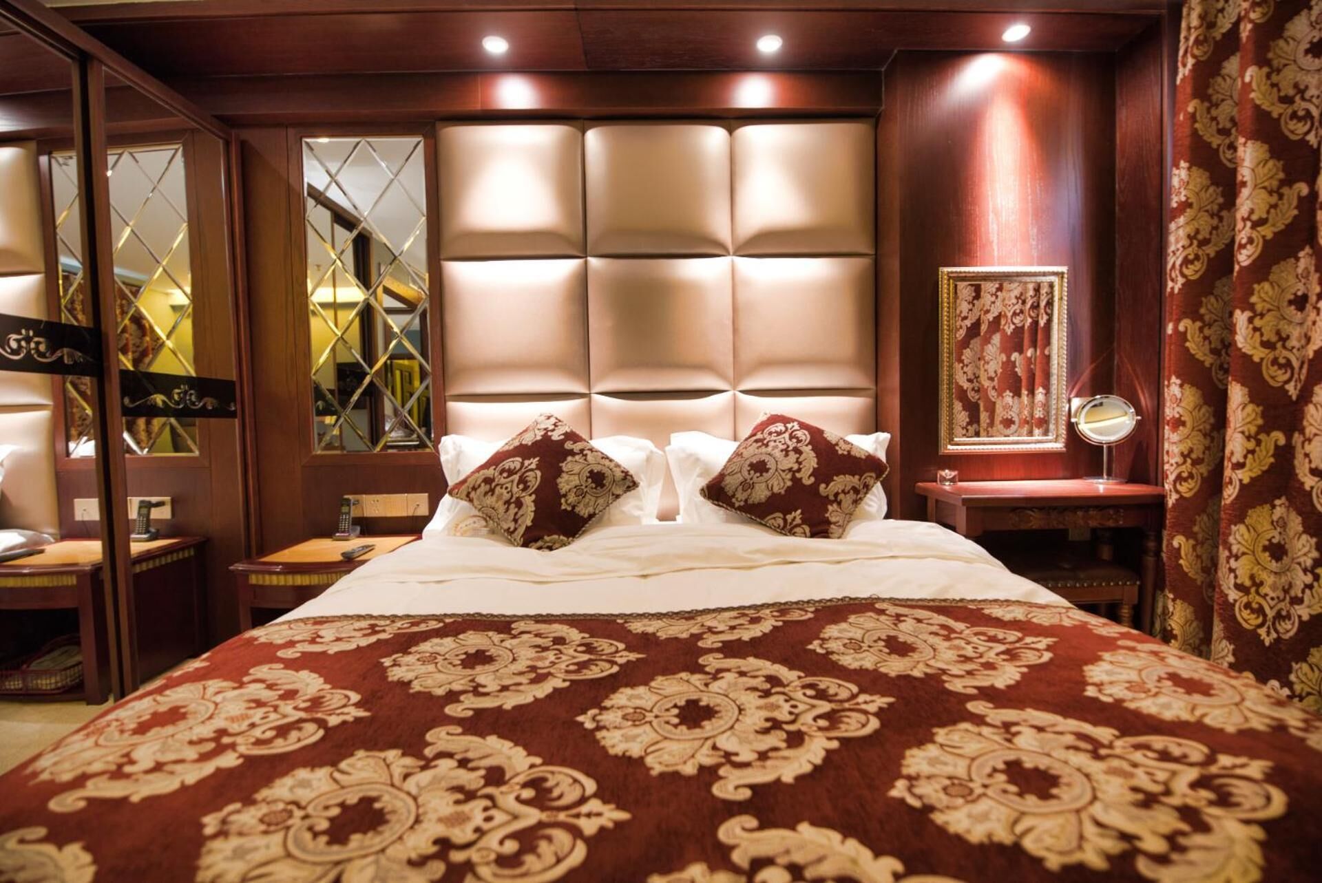 A hotel room with a large bed, a headboard with a light panel, a bedside table with a mirror, and a patterned carpet.
