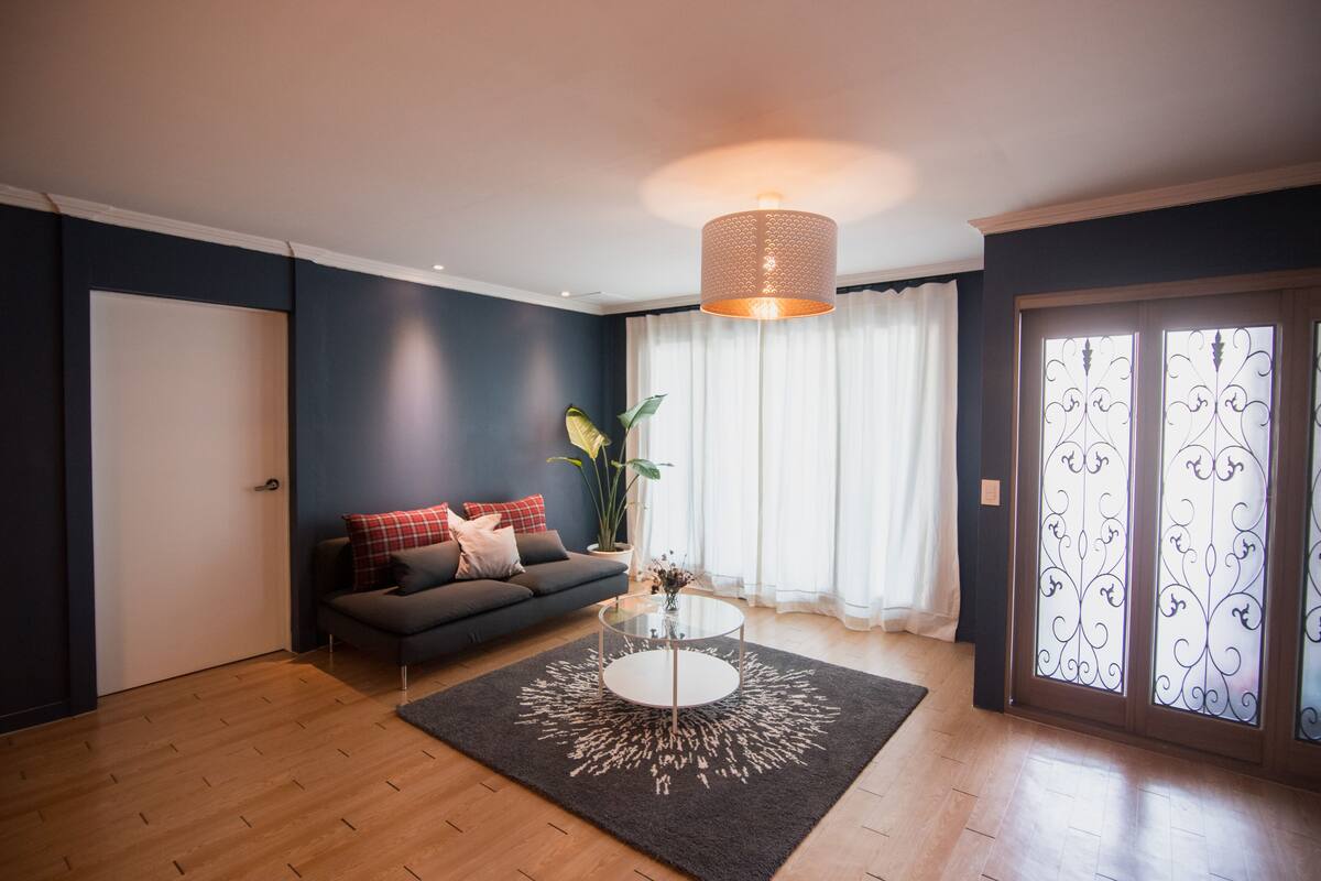 Ein Wohnzimmer mit dunkelblauer Wand, einem grauen Sofa mit roten und weißen Kissen, einem Glastisch und einer Hängeleuchte.