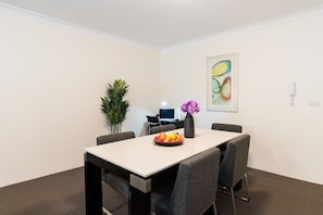 A conference room with a white table, four grey chairs, a vase with purple flowers, and a fruit bowl. There is a potted plant, a desk with a computer, and a colorful abstract painting on the wall.
