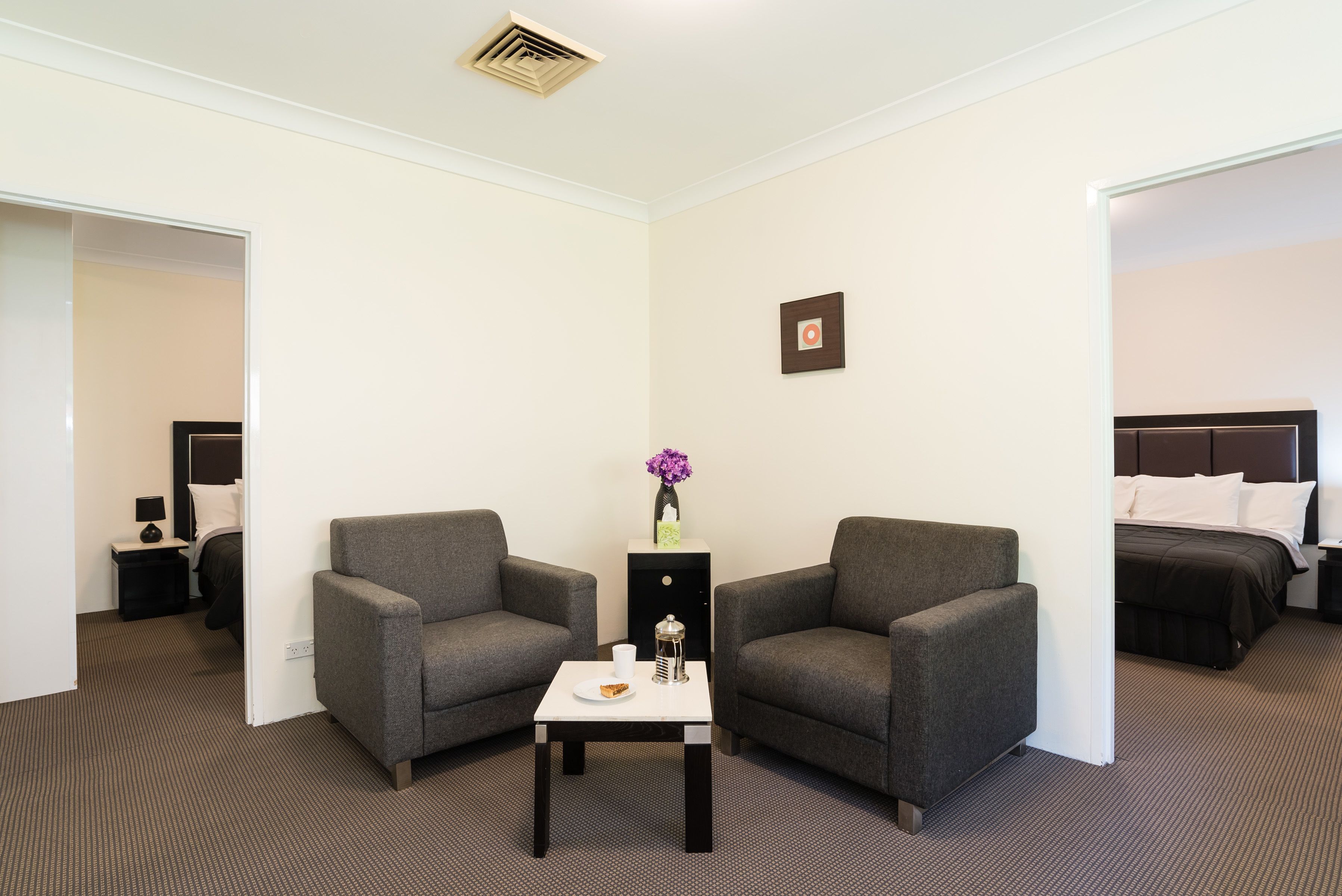 Habitación de hotel con dos sillones grises, una mesita con una taza y un plato pequeño, una mesita de noche con una lámpara, y una cama con ropa blanca.