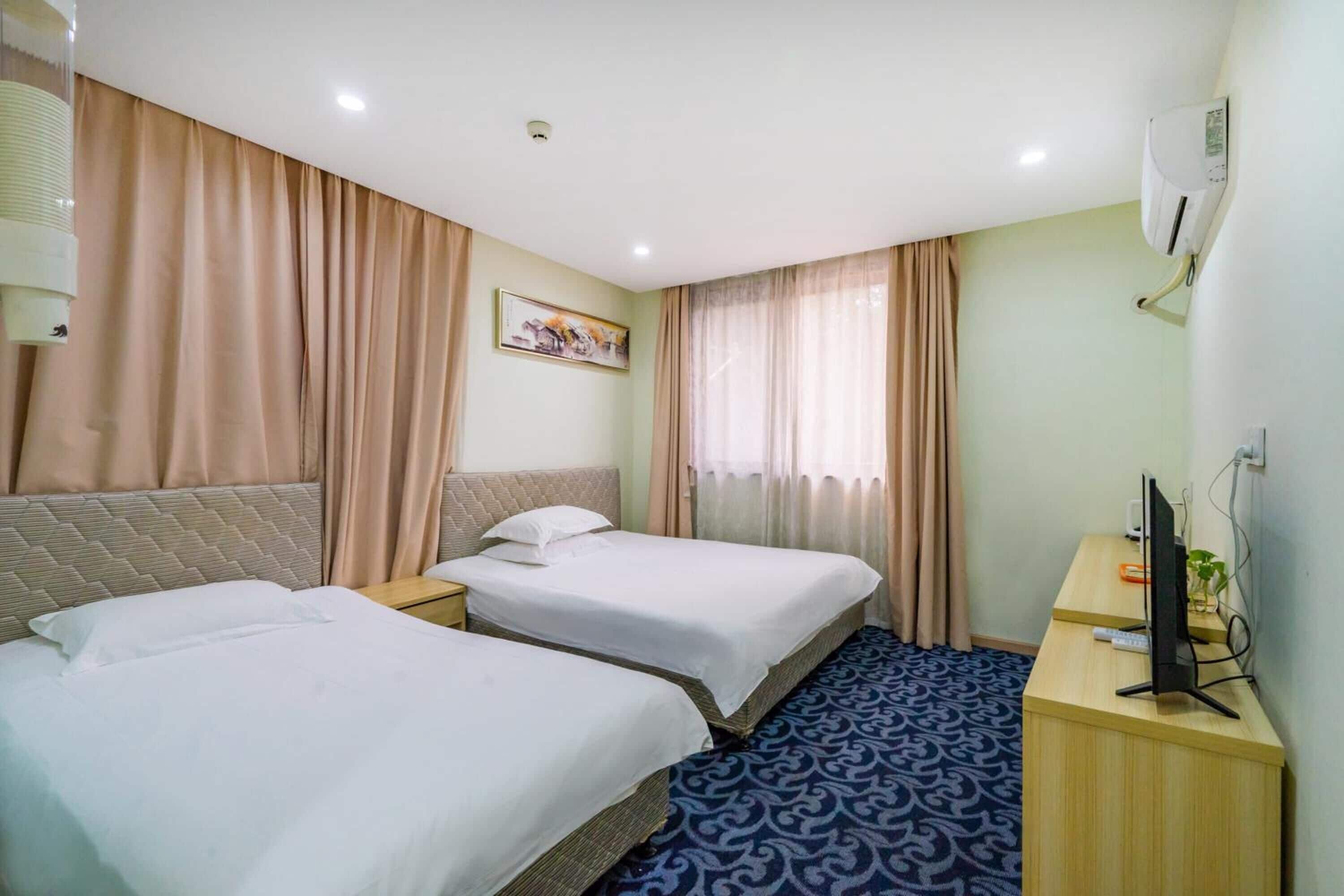 A hotel room with two beds, a desk with a computer, a wall-mounted air conditioner, and a window with curtains.