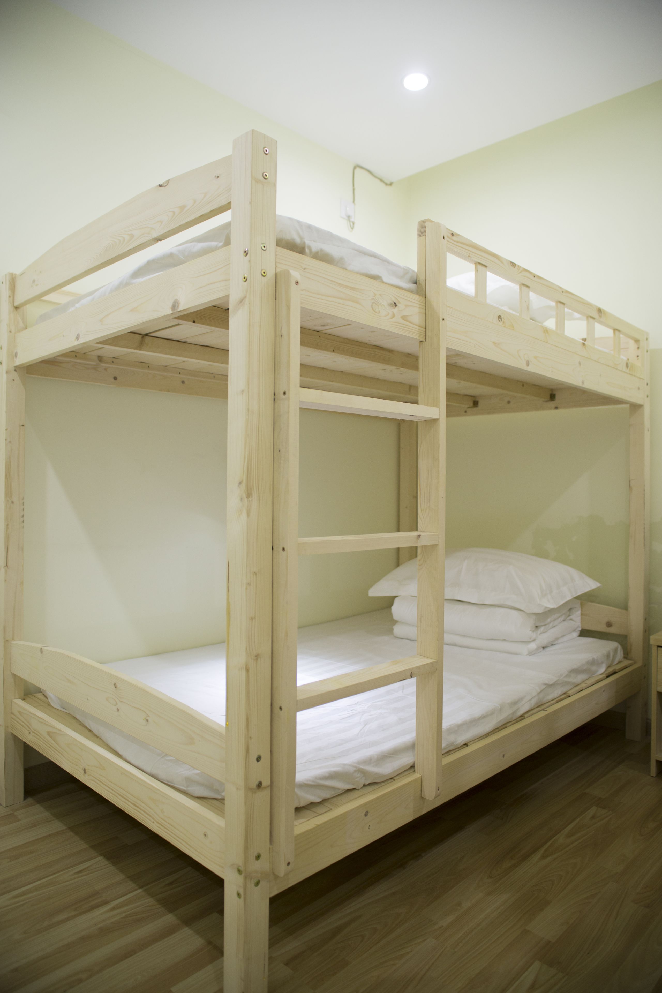 A bunk bed with a wooden frame and white bedding, in a room with light-colored walls and wooden flooring.