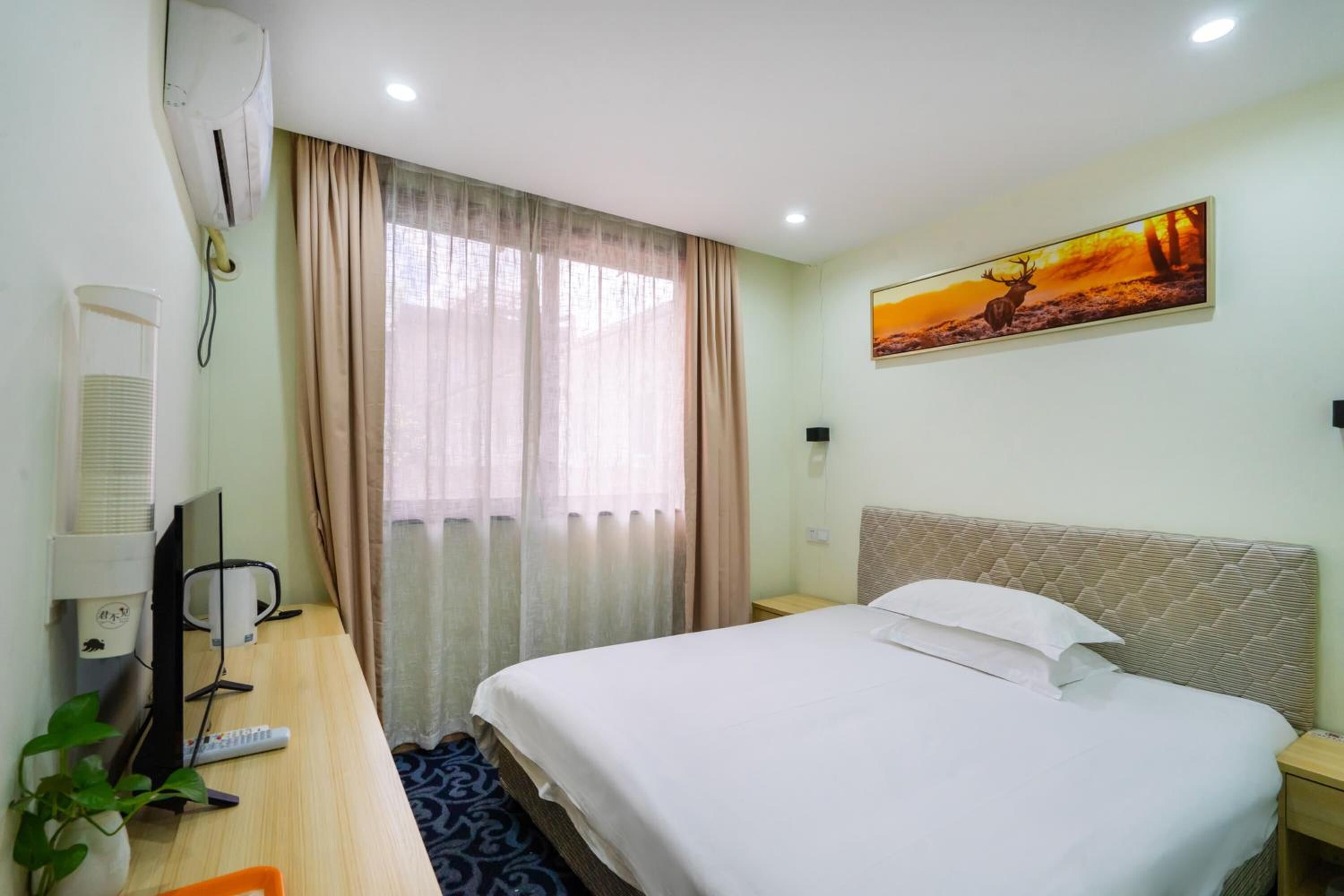 A hotel room with a bed, a desk with a computer and a kettle, a wall-mounted air conditioner, a window with curtains, and a painting on the wall.