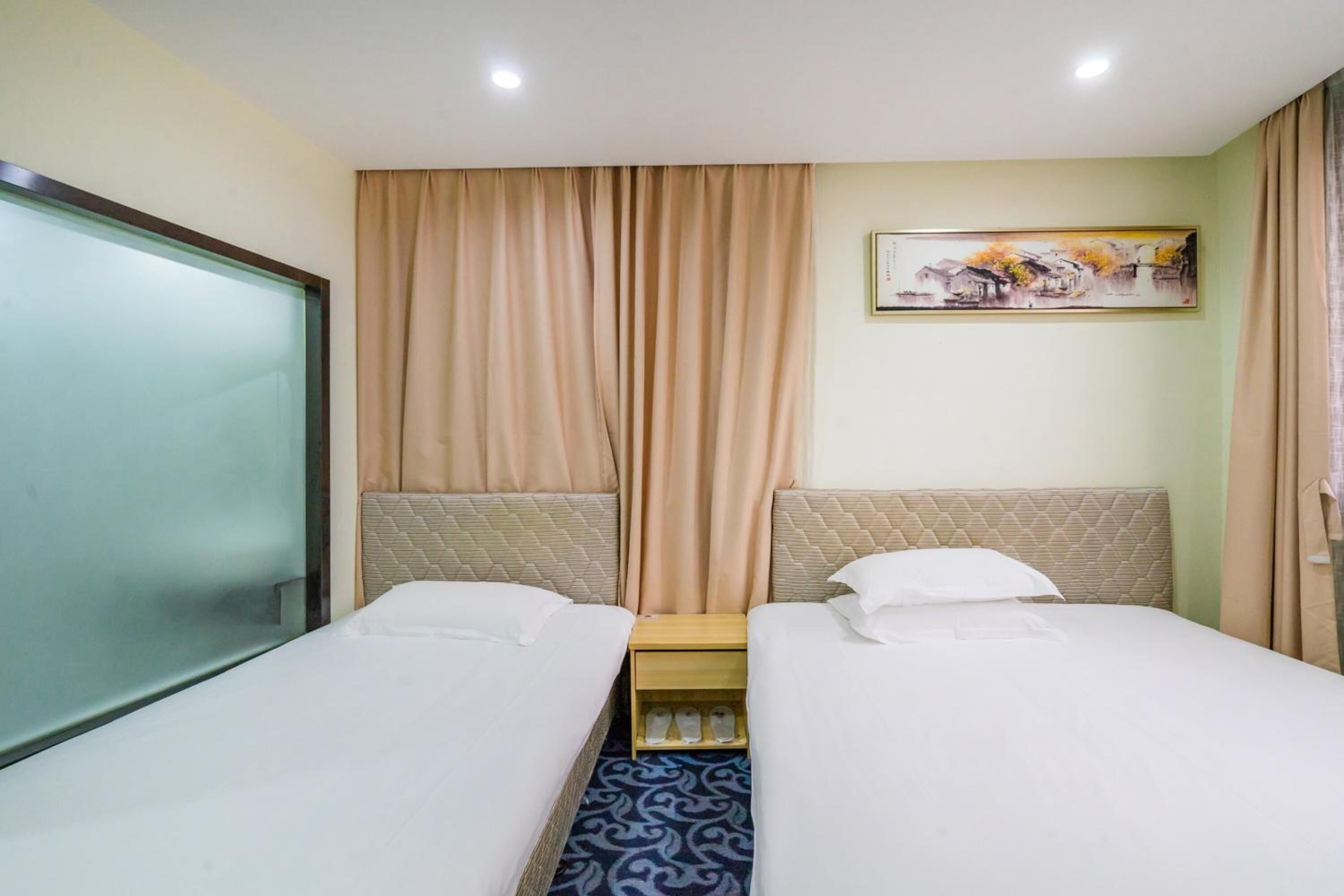 Habitación de hotel con dos camas, cortinas color beige, un cuadro en la pared y una mesita de noche de madera.