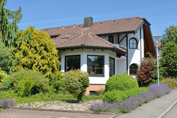 A two-story house with a tiled roof, surrounded by lush greenery and flowering plants.