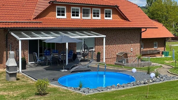 Uma casa de dois andares com telhado de telhas vermelhas, um alpendre com mobiliário para área externa e uma piscina.